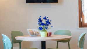 Una mesa redonda con mantel blanco, dos sillas verdes, una planta en maceta con flores azules y una cesta tejida blanca.