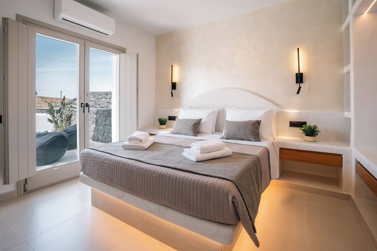 A modern bedroom with a large bed, bedside tables, wall-mounted lights, and a view through a sliding door.