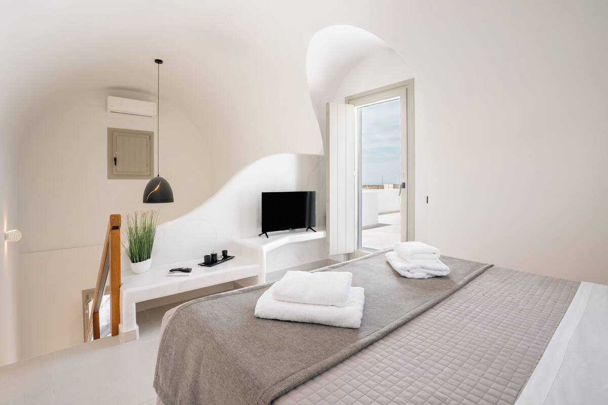 A modern bedroom with a bed, a TV, a desk, and a view of the sky.