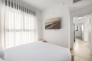 Ein Hotelzimmer mit Bett, einem an der Wand befestigten Fernseher und Blick auf den Strand durch das Fenster.