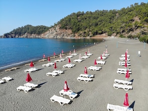 A beach with rows of white lounge chairs and red umbrellas, a clear blue sky, and a forested hillside in the background. - GÜNLÜ HOTEL (Fethiye)