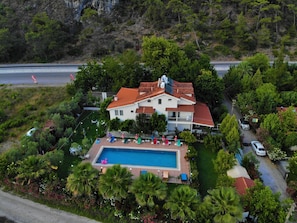 A two-story house with a swimming pool, surrounded by lush greenery and a mountainous backdrop.