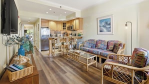 A living room with a sofa, chairs, a coffee table, a TV, and a kitchen area with cabinets and appliances. - Dunes Day at Villas of Hatteras Landing by KEES Vacations (Hatteras)