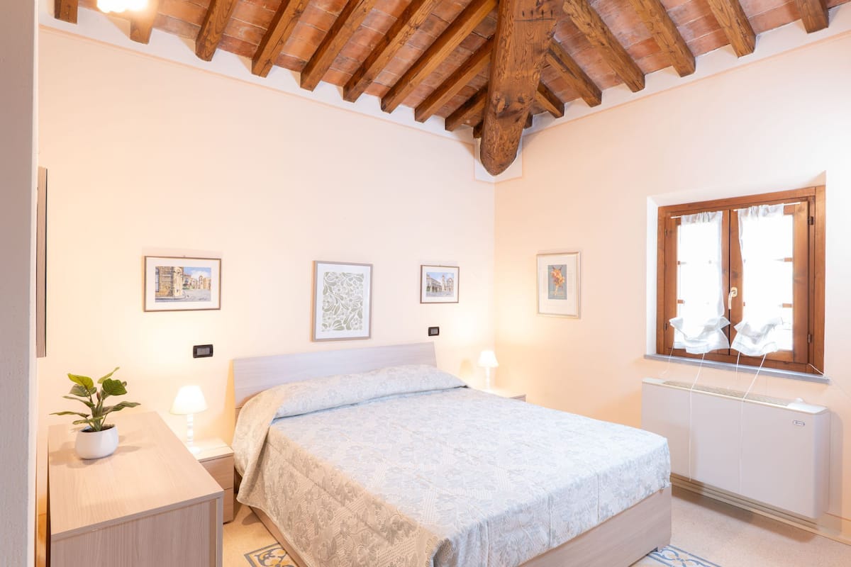 A bedroom with a wooden ceiling, a bed, a bedside table with a lamp, a radiator, and framed pictures on the wall.