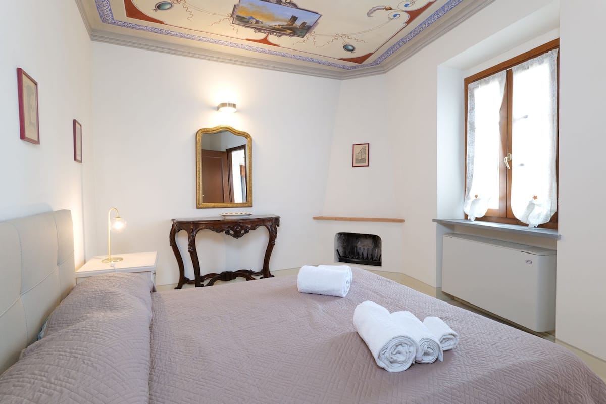 A hotel room with a bed, a bedside table with a lamp, a mirror, a window with curtains, and a radiator.