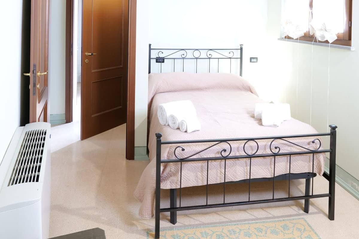 A bedroom with a bed, wooden ceiling beams, a radiator, a window with curtains, and a door.