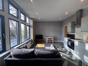 Uma sala de estar moderna com um sofá, TV e uma pequena mesa. Uma cozinha com micro-ondas e forno é visível ao fundo.