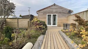 Una casa de madera con puerta principal, un porche pequeño y un jardín con plantas y una terraza de madera.