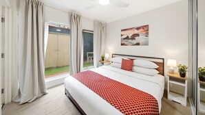 Una habitación de hotel con una cama, un ventilador de techo, un ventanal con cortinas, un cuadro en la pared y una mesita de noche con una lámpara y una planta.