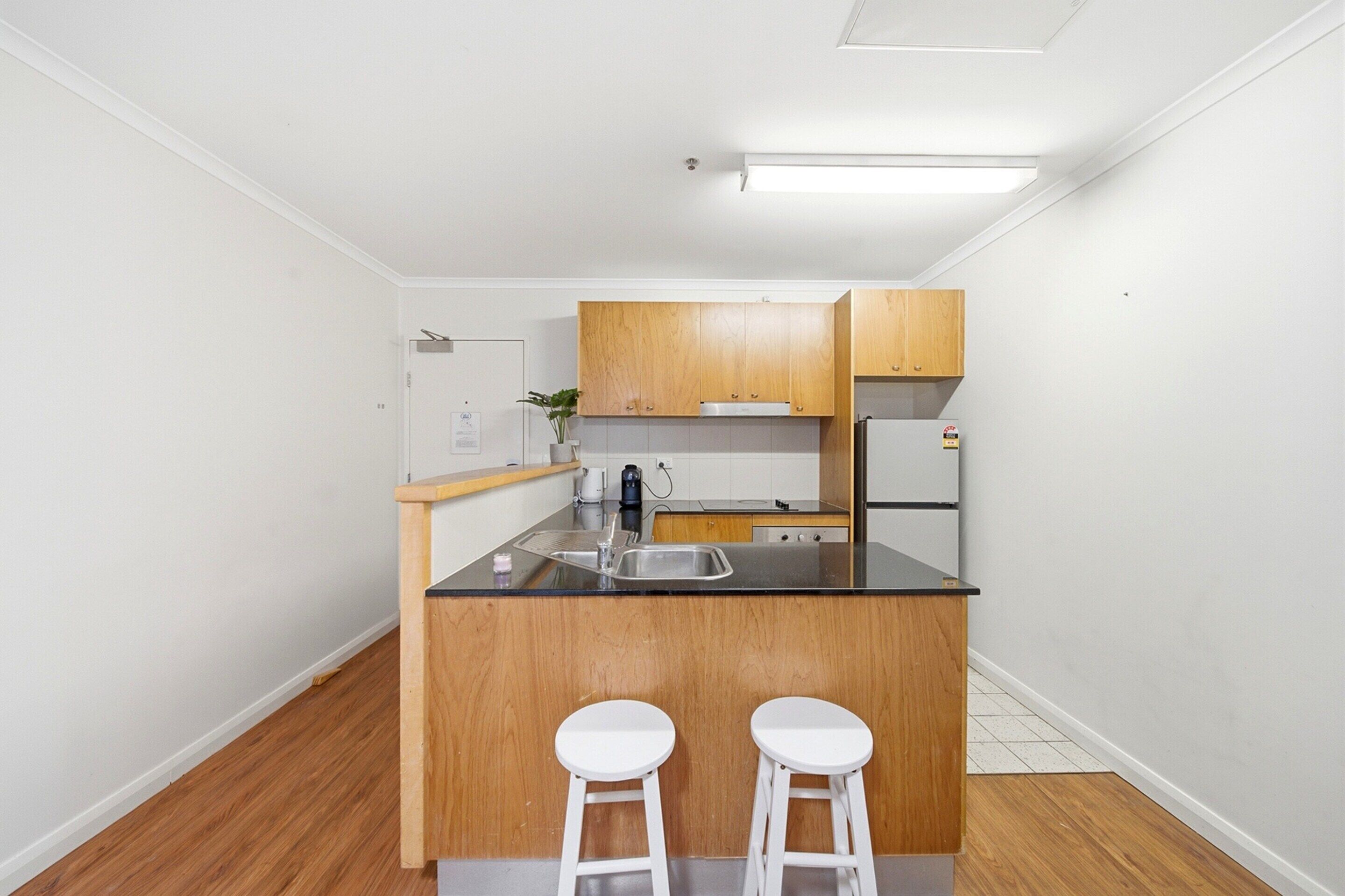 A compact kitchen with wooden cabinets, a black countertop, a sink, and two white stools.