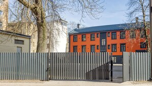 Ein verschlossenes Tor mit einem Metallzaun, ein Gebäude mit roten und schwarzen Außenwänden und ein Baum im Vordergrund.