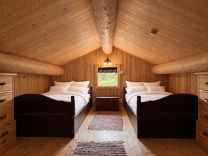 A room with wooden walls and ceiling, two beds with white bedding, a small window with a view, and a wooden dresser with drawers.