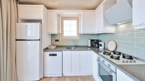 A compact kitchen with white cabinetry, a built-in dishwasher, a stove with four burners, and a window above the sink. - AURELİS SUİTES (KAŞ)