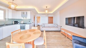 A modern kitchen with white cabinets, a dining table with chairs, a sofa, a TV, and a bench. - AURELİS SUİTES (KAŞ)