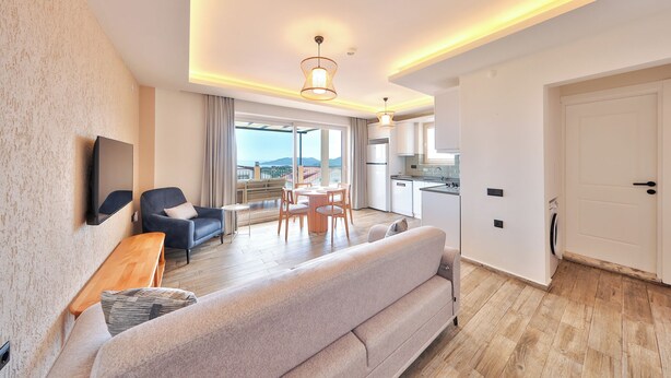 A modern living room with a sofa, a dining table, and chairs, a TV mounted on the wall, and a kitchen area with a white door. - AURELİS SUİTES (KAŞ)
