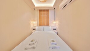 A hotel room with a bed, two bedside tables, a ceiling light fixture, a window with blinds, and an air conditioning unit. - AURELİS SUİTES (KAŞ)