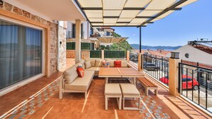 A balcony with a sofa, table, and chairs, overlooking a street with cars and buildings. - AURELİS SUİTES (KAŞ)
