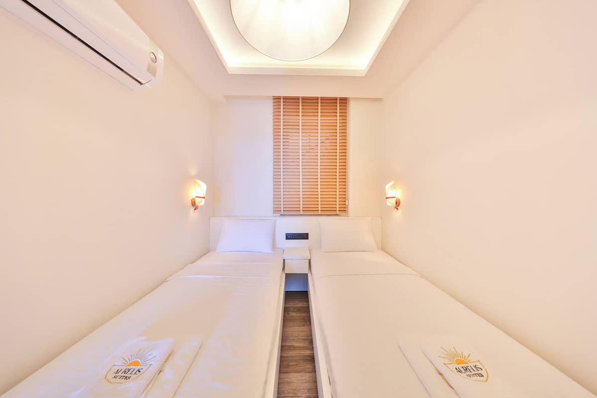 Une chambre d’hôtel avec deux lits, un luminaire au plafond, une fenêtre avec des stores et des lampes fixées au mur.