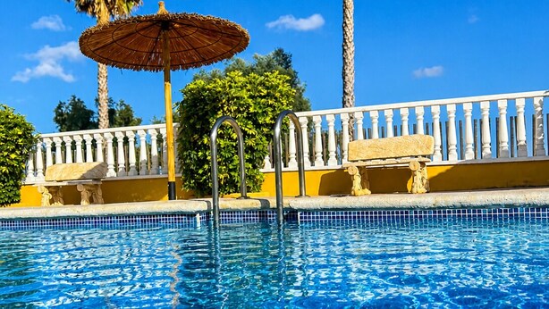 Una piscina con agua azul cristalina, rodeada por una barandilla blanca, palmeras y un paraguas de paja.