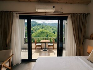 A bedroom with a bed, a window with curtains, a view of green hills, and a balcony with a table and chairs. - Jakuzili Deluxe Suite • Vadi Manzaralı • Ardeşen (Rize)