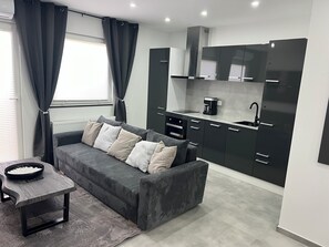 Ein modernes Wohnzimmer mit einer Couch und einem Couchtisch, eine KĂŒche mit dunklen SchrĂ€nken und einem eingebauten Backofen sowie ein Fenster mit VorhĂ€ngen.