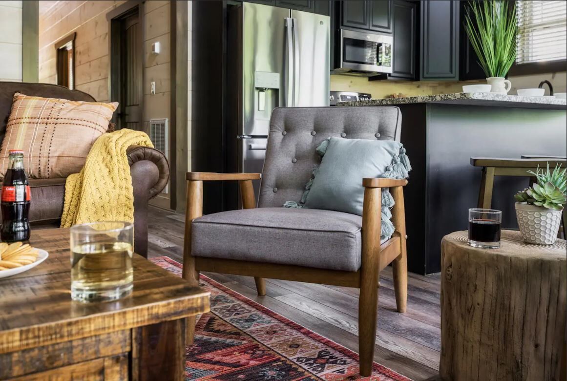 A modern living room with a grey upholstered armchair, a wooden coffee table, and a kitchen area with dark cabinetry and a microwave.