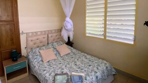 Une chambre à coucher comprenant un lit, une table de chevet, une commode, une fenêtre avec des stores et un ventilateur de plafond.