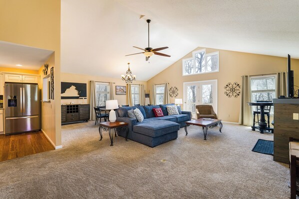 A spacious living room with a blue sectional sofa, a chandelier, a ceiling fan, and large windows.