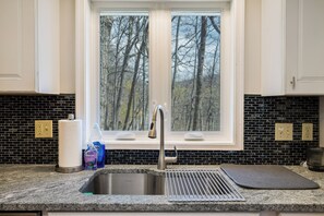 A kitchen with a stainless steel sink, a window with a view of trees, and a black and white tiled backsplash.