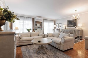 Ein Wohnzimmer mit einem beige gefärbten Sofagarnitur, einem runden Couchtisch, einem gemusterten Teppich, einem Bücherregal, einem Gemälde und einer Chandelier.