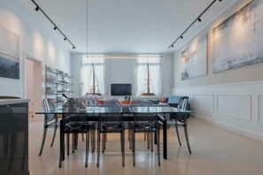 Een moderne eethoek met een glazen tafel, stoelen, een televisie en een boekenplank.