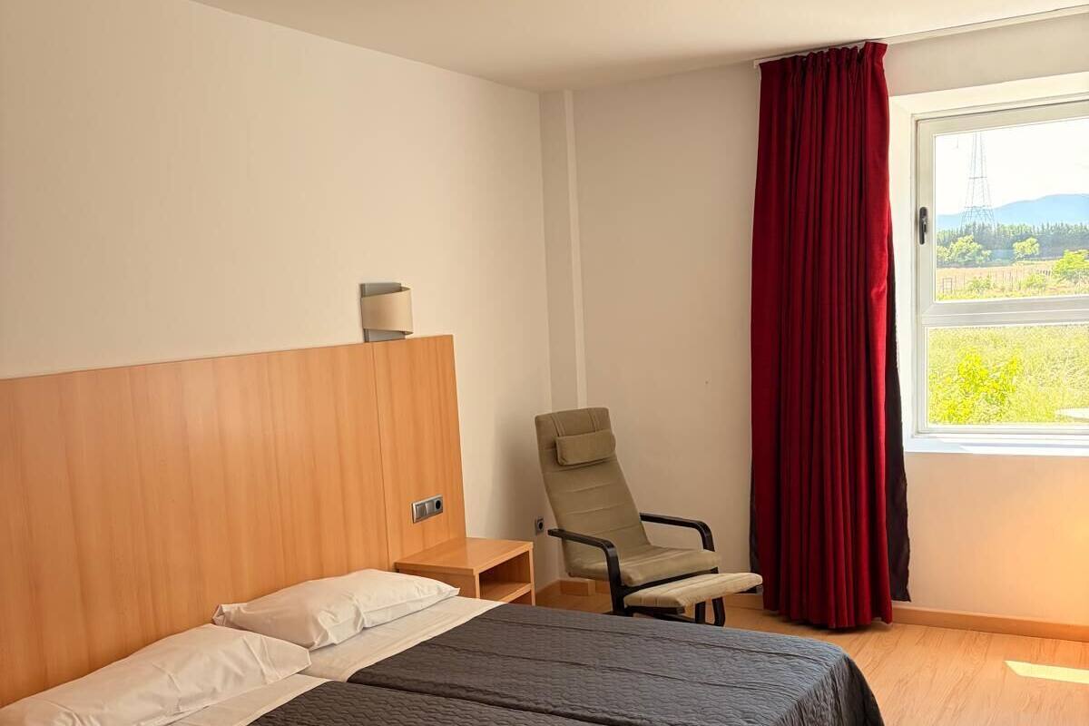Una habitación de hotel con cama, una silla, un ventanal y un equipo instalado en el techo.