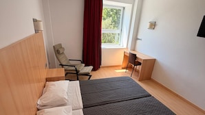 Quarto de hotel com duas camas, uma escrivaninha, uma cadeira, uma janela com cortinas vermelhas e uma TV fixada na parede.
