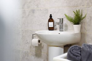 A bathroom with a white sink, a bottle of hand soap, a faucet, a roll of toilet paper, and a potted plant. - Clarence Cottage | 4mins to Pier, Pet Friendly with Shared Garden (Portsmouth)