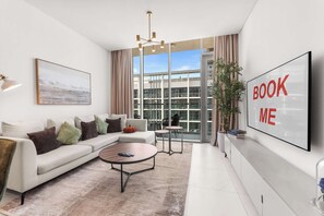 Ein modernes Wohnzimmer mit einer Eckcouch, einem Couchtisch, einem Fernseher und einem großen Fenster mit Blick auf ein Gebäude.