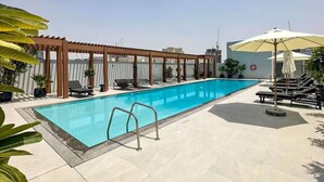 Ein rechteckiger Swimmingpool mit einer Holzpergola und Liegestühlen an einer Seite sowie ein überdachter Bereich mit Sonnenschirmen auf der anderen Seite.