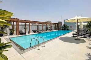A rectangular swimming pool with a wooden pergola and lounge chairs on one side, and a covered area with umbrellas on the other. - Vogue, JVC Luma22 West near Circle Mall (Jumeirah Village Circle ( JVC ))