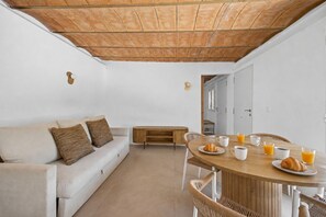 Une chambre moderne avec un plafond en bois, des murs blancs, un canapé avec des coussins marron, une table à manger en bois avec des chaises, et des éléments pour le petit-déjeuner.