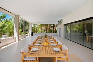 A spacious dining area with a long wooden table set for a meal, surrounded by chairs, and large windows offering a view of greenery.