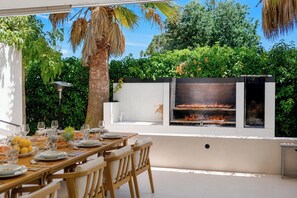 Un espace extérieur aménagé pour les repas, avec un barbecue, des murs blancs et de la verdure.