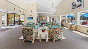 A spacious lobby with teal sofas, a central white table, and large windows with blue umbrellas. - Coastal Getaway Arcadian Dunes #250 -Steps to Sand (Myrtle Beach)