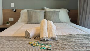 Ein ordentlich bezogenes Bett mit zusammengerollten Handtüchern und einer Decke, ein Kopfteil, ein Nachttisch mit einer kleinen Pflanze und drei gerahmte Bilder an der Wand.