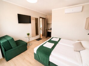 A hotel room with a bed, a green sofa, a small table with a plant, a TV mounted on the wall, and a kitchen area visible through an open door. - Apartment Near the Sea, Breakfast Included (Dziwnów)