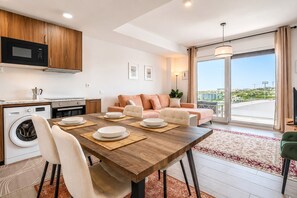 Eine moderne Küche mit Mikrowelle, Backofen und Waschmaschine; ein Essbereich mit Holztisch und Stühlen; ein Sofa mit Kissen; und eine große Glasschiebetür mit Blick auf die Stadt.