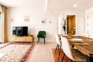 Ein modernes Wohnzimmer mit einem Flachbildfernseher, einem grünen Sessel, einem Esstisch mit Stühlen und einem Teppich mit roten und weißen Mustern.