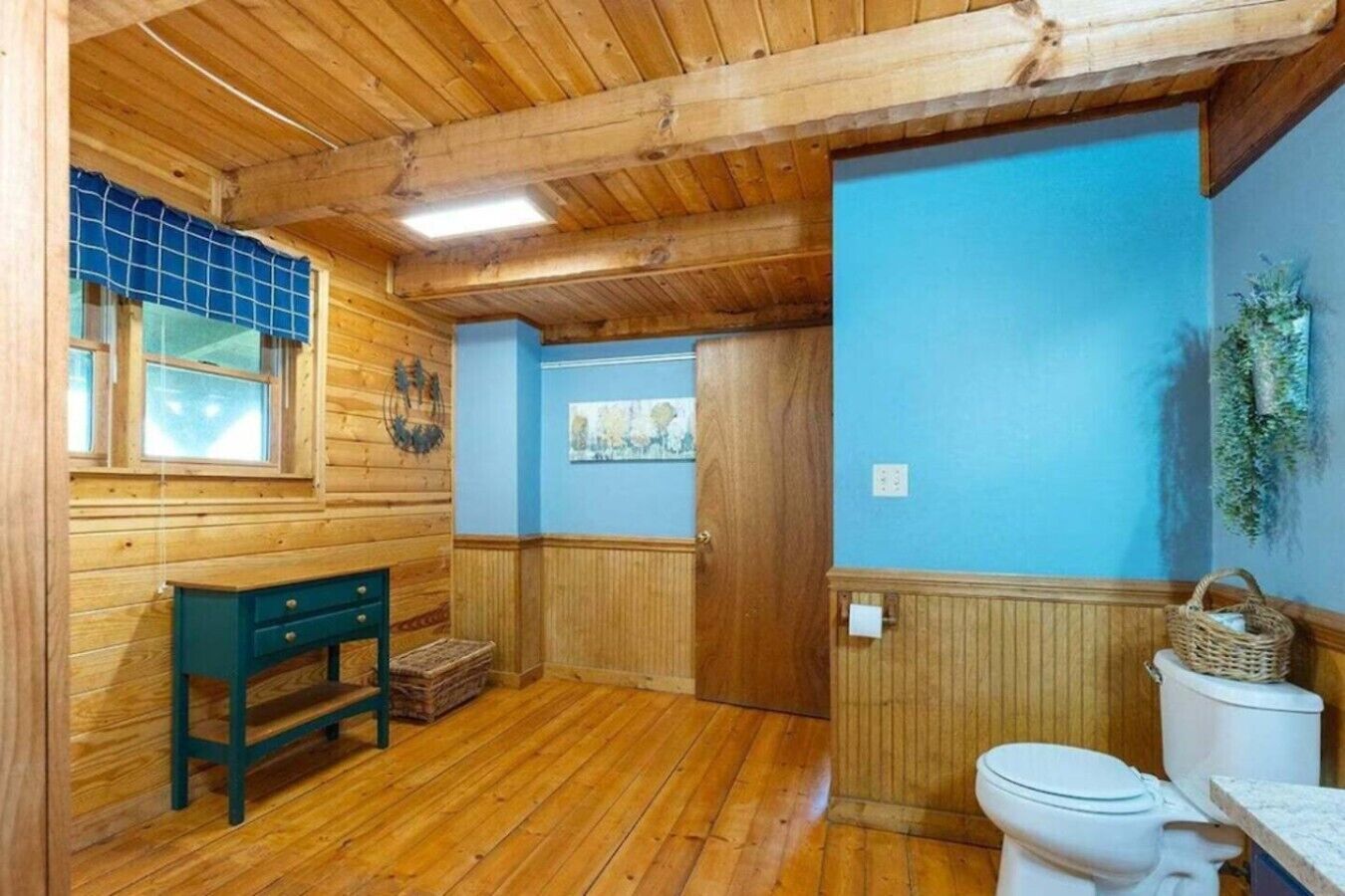 L’intérieur d’une cabane en bois, avec un plancher en bois, un mur bleu, un banc en bois, des toilettes blanches et une fenêtre dotée de vitres teintées en bleu.