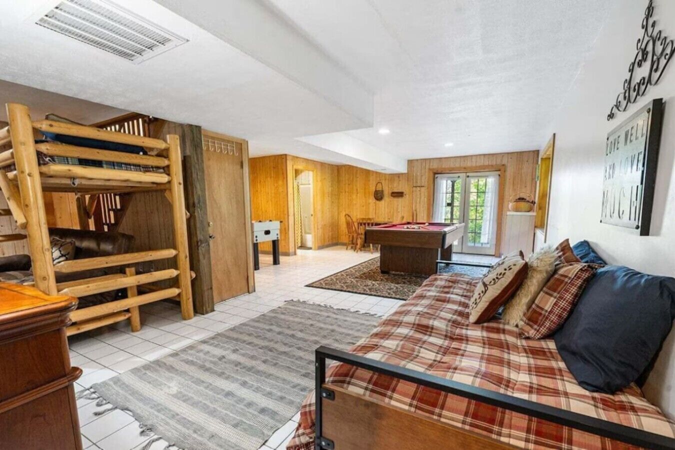 Une pièce avec une table de billard, un canapé avec des coussins à carreaux, une structure de jeu en bois et une porte donnant sur une autre pièce.