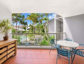 Un balcon avec vue sur les palmiers et une piscine, une table en bois et des chaises bleues.