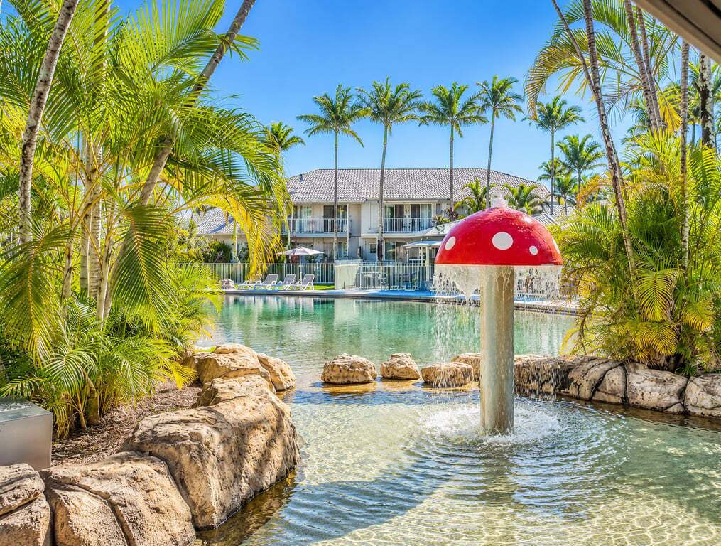 A resort pool with a mushroom-shaped fountain, palm trees, and a building in the background.