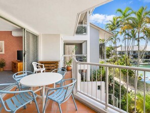 A balcony with a table and chairs, a TV, and a view of a pool and palm trees. - Modern Mermaid Beach | Resort Pool & Walk to Beac (Gold Coast)
